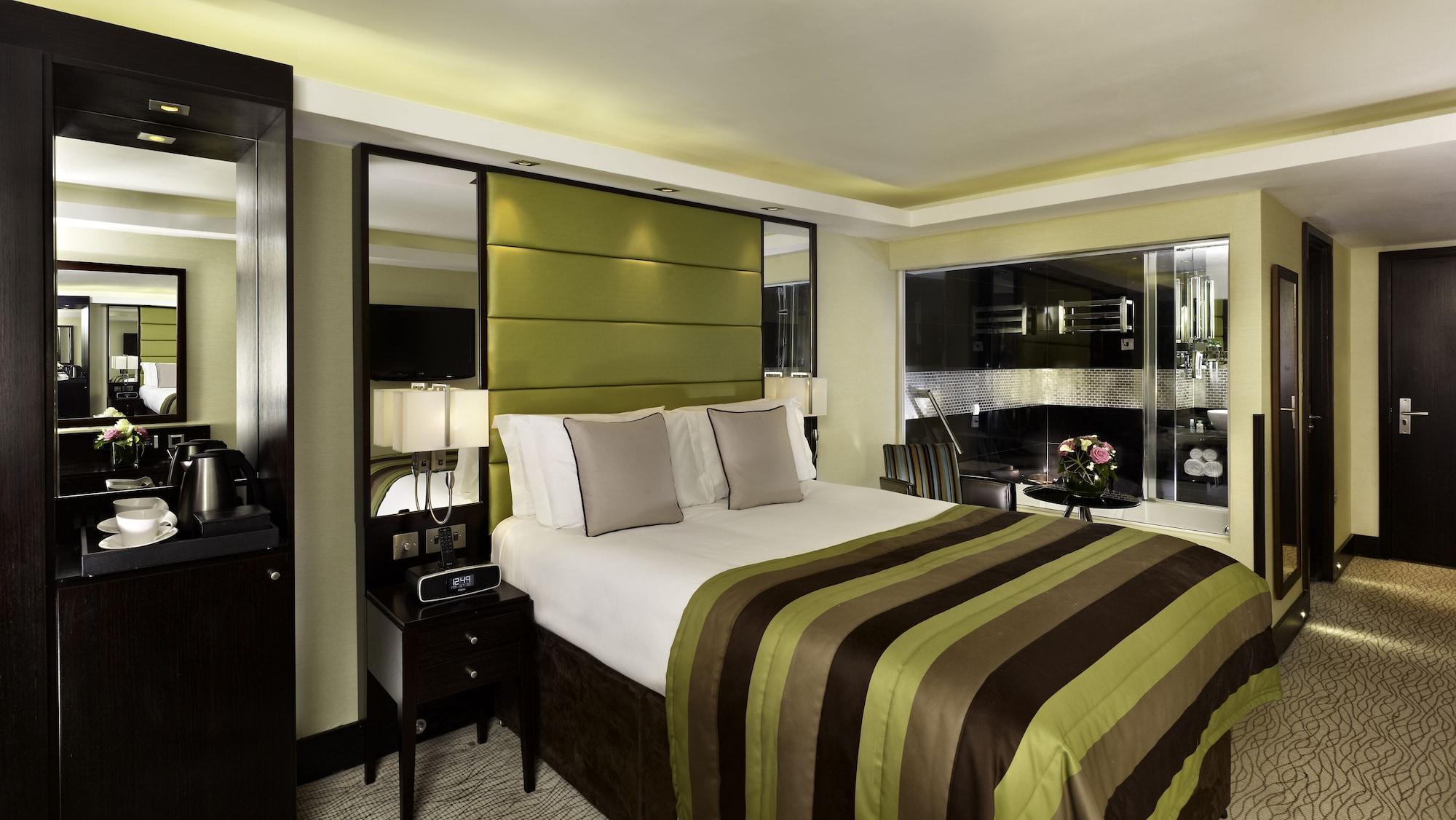 Отель Montcalm Brewery Townhouse Лондон Экстерьер фото The photo shows a modern hotel room designed with a sophisticated aesthetic. It features a comfortable double bed with a striped brown and green bedspread, complemented by two light gray decorative pillows. Behind the bed is an elegant headboard sect
