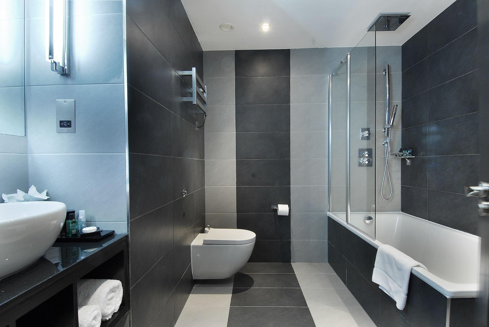 Отель Montcalm Brewery Townhouse Лондон Экстерьер фото The photo shows a modern bathroom featuring a sleek design. The walls are tiled in a combination of dark gray and light gray, creating a contemporary look. On the left, there is a countertop with a basin and some toiletries placed on it. The middle s