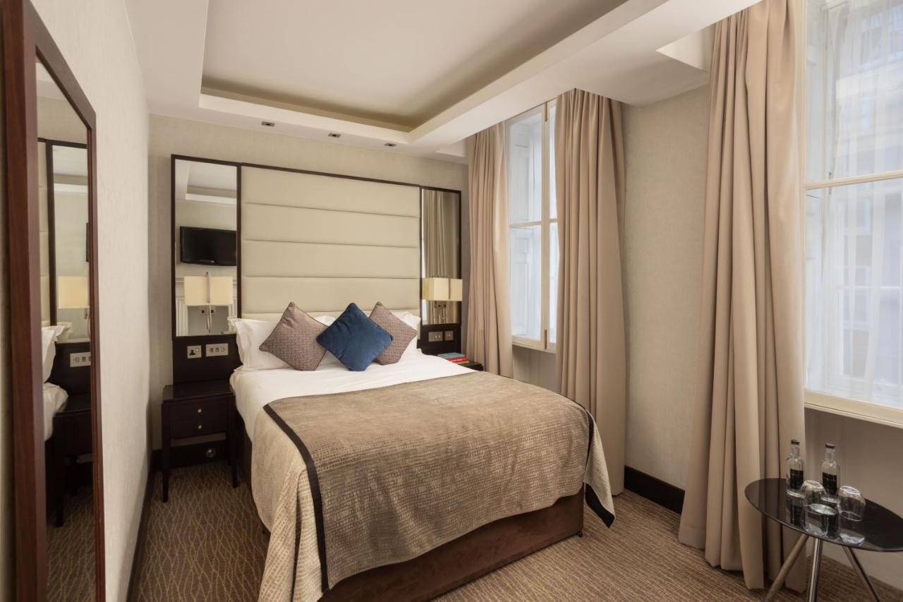 Отель Montcalm Brewery Townhouse Лондон Экстерьер фото The photo shows a cozy hotel bedroom. The room features a neatly made double bed with a beige bedspread and decorative pillows, including a blue one. There are bedside tables on either side of the bed with lamps. The walls are painted in a soft color