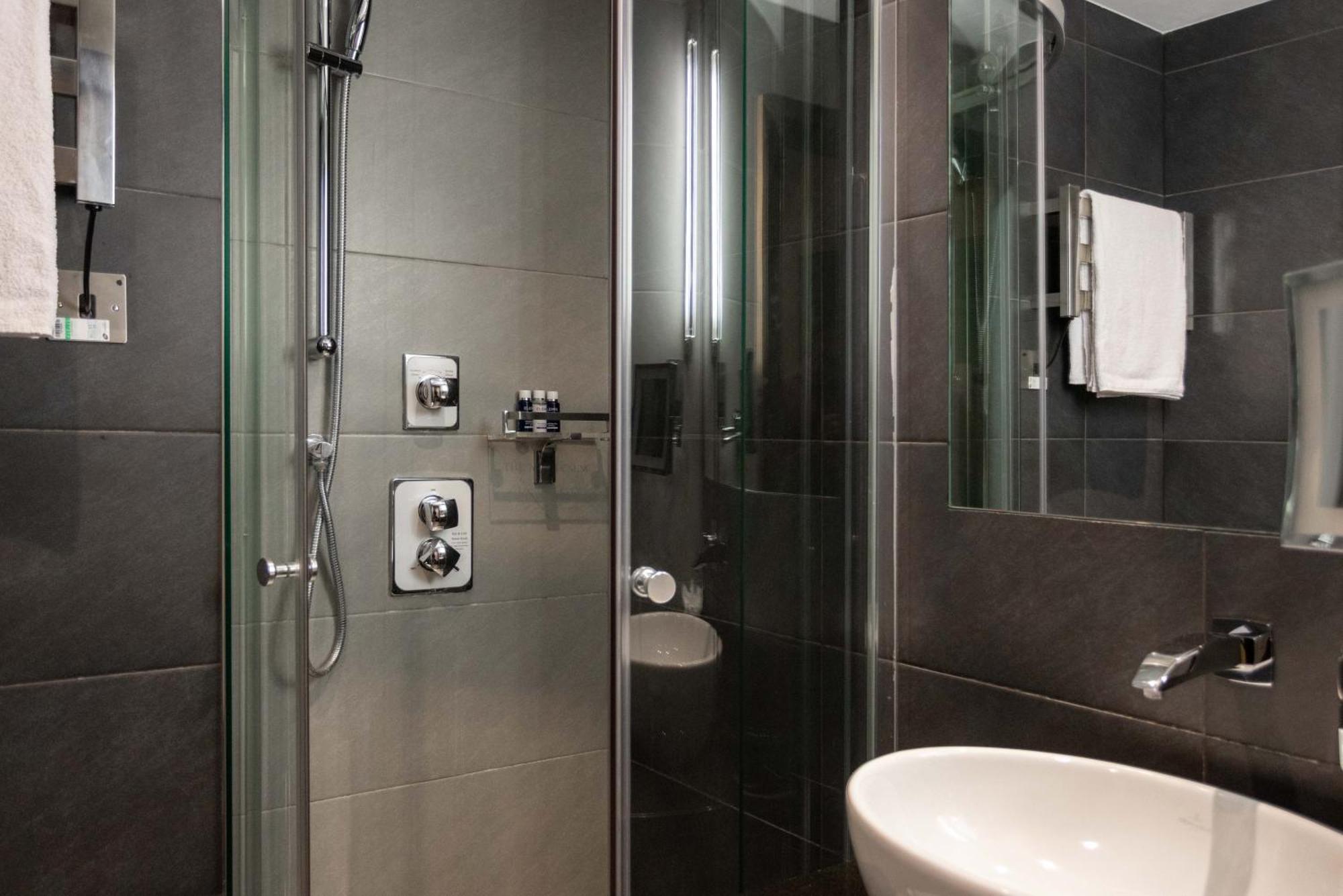 Отель Montcalm Brewery Townhouse Лондон Экстерьер фото The photo shows a modern bathroom with a glass shower enclosure. The shower features a handheld showerhead and various controls mounted on the wall. The walls are tiled in dark, sleek tones, contributing to a contemporary aesthetic. There is a round 