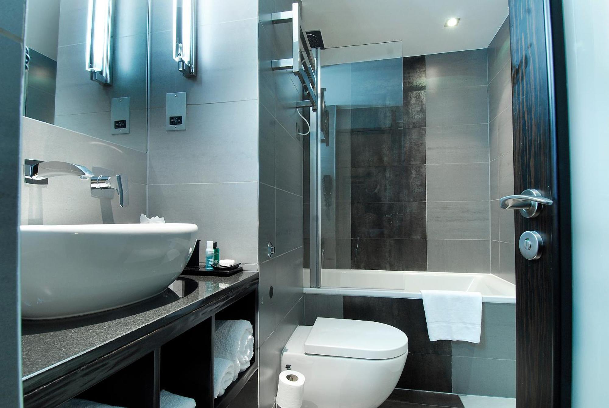 Отель Montcalm Brewery Townhouse Лондон Экстерьер фото The photo shows a modern bathroom with sleek design elements. It features a curved white sink on a dark countertop, with a mirror above that is illuminated by stylish lighting fixtures. Next to the sink, there's a toilet, and on the opposite side, a 