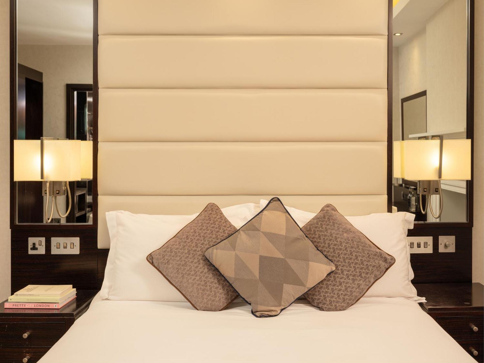Отель Montcalm Brewery Townhouse Лондон Экстерьер фото The image shows a cozy bedroom setting. The bed features a headboard upholstered in a light beige fabric. It is adorned with three decorative pillows in varying patterns and colors, placed on white bedding. On either side of the bed, there are modern