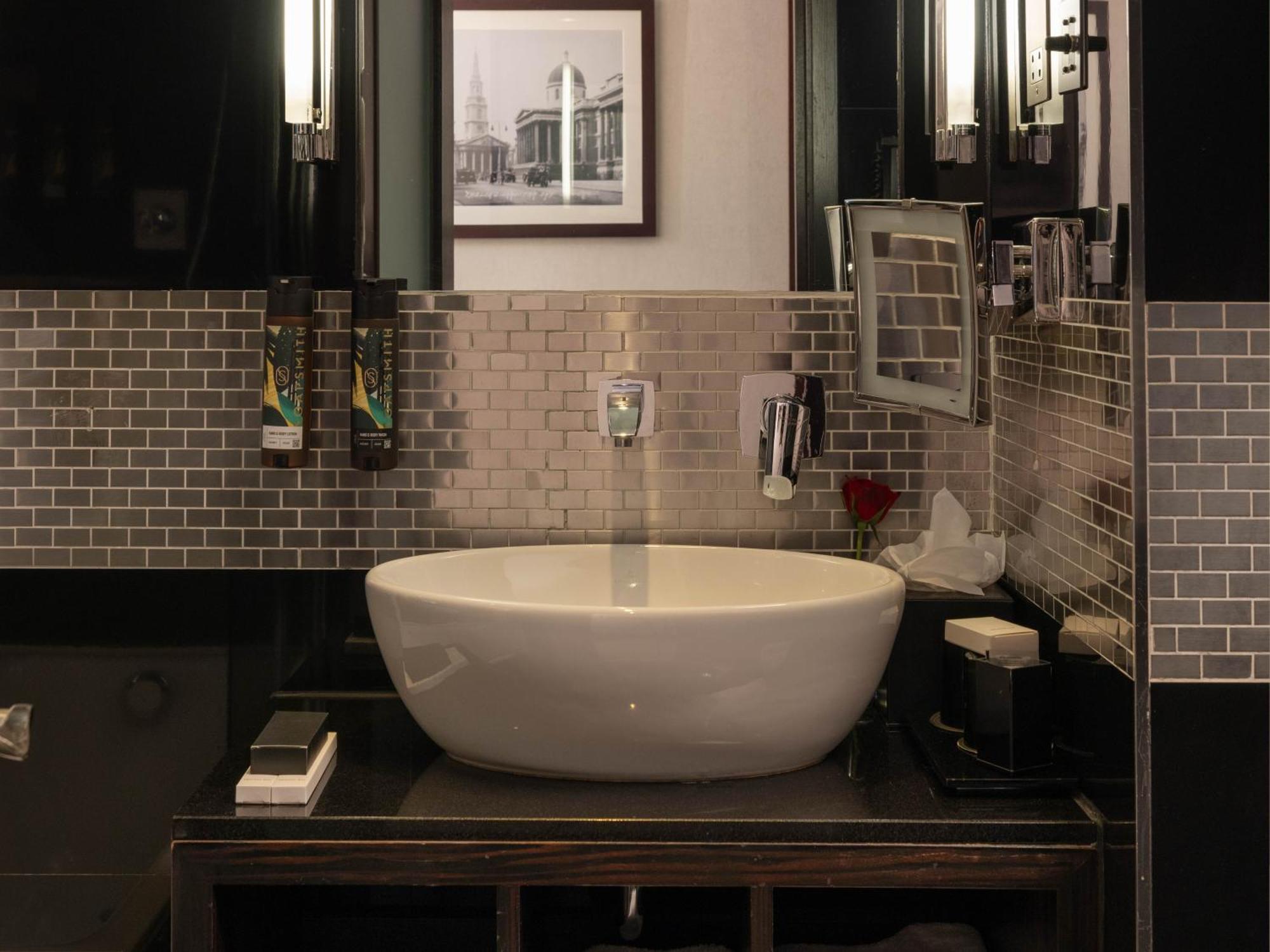 Отель Montcalm Brewery Townhouse Лондон Экстерьер фото The photo shows a modern bathroom with a stylish design. It features a large, round white sink placed on a dark wooden vanity. The walls are adorned with a mix of gray and silver tile, creating a sleek and contemporary look. Above the sink, there is 
