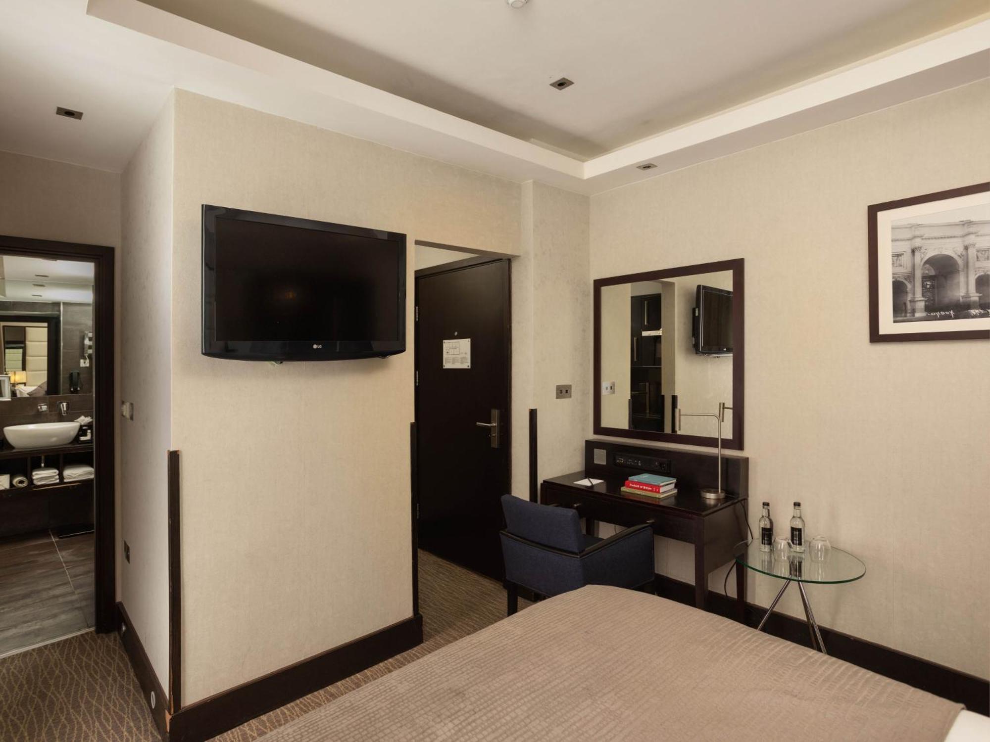 Отель Montcalm Brewery Townhouse Лондон Экстерьер фото The photo shows a hotel room with a modern and minimalist design. On the left, there's a wall-mounted flat-screen TV. In the center, there's a bed with a neutral-colored bedspread. To the right of the bed, there is a small desk with a mirror above it