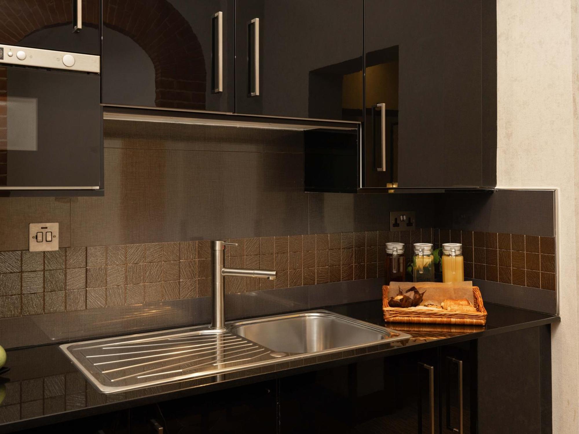 Отель Montcalm Brewery Townhouse Лондон Экстерьер фото The photo shows a modern kitchen with a sleek design. It features dark cabinetry with a glossy finish and stainless steel handles. There is a stainless steel sink with a simple faucet, and a countertop made of black material. Above the sink, there ar
