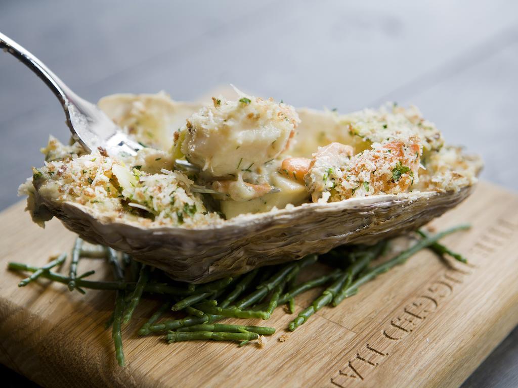 Отель Montcalm Brewery Townhouse Лондон Экстерьер фото The photo shows a large oyster shell filled with a mixture that appears to include seafood, herbs, and breadcrumbs. The dish is presented on a wooden board with some greenery, possibly a type of seaweed or garnishing. A fork is placed beside the oyst