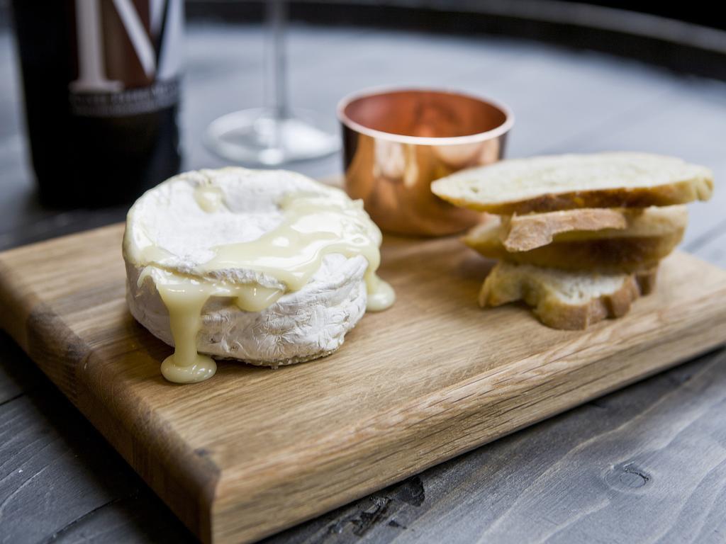 Отель Montcalm Brewery Townhouse Лондон Экстерьер фото The photo features a wooden serving board with several items. There is a round cheese, likely Brie or a similar soft cheese, that appears to be melting and dripping slightly. Next to the cheese, there is a small copper cup that might contain a sauce 