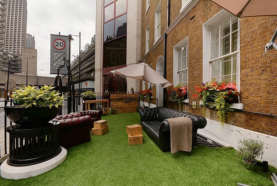 Отель Montcalm Brewery Townhouse Лондон Экстерьер фото The photo depicts a charming outdoor space, likely a patio or garden area. It features two comfortable-looking leather sofas, one of which is a deep burgundy color, placed on a green artificial lawn. There are wooden crates that could serve as side t